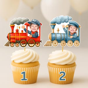 May include: Two cupcakes topped with personalized train-themed cake toppers. One topper features a red steam engine with a child's photo, the other a blue steam engine with a child's photo. Both trains have smoke clouds and are labeled 1 and 2.