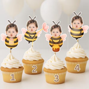 May include: Four cupcakes with white icing, each adorned with a bee-themed topper featuring a child's face. The bee decorations have black and yellow stripes, pink wings, and hold honey pots or flowers. The cupcakes are numbered 1 to 4.