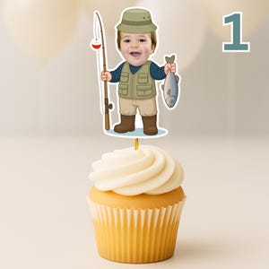 K&ouml;nnte beinhalten: Ein Cupcake mit einem dekorativen Papieraufstecker, der eine Comic-Illustration eines Kindes mit beigem Anglerhut, blauem Langarmshirt, beiger Anglerweste mit Taschen, beiger Hose und braunen Stiefeln zeigt, das eine Angel und einen Fisch h&auml;lt.