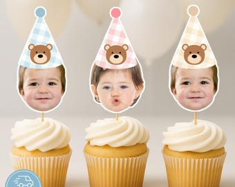 Puede incluir: Tres cupcakes con glaseado blanco, cada uno coronado con una foto de un niño con un gorro de fiesta con un diseño de oso de peluche. Los sombreros son de color azul claro, rosa y beige con un patrón de cuadros. Los cupcakes están sobre una superficie blanca.