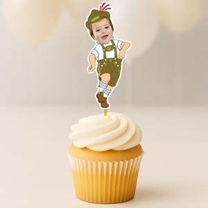 May include: A cupcake topper featuring a cutout of a child's face attached to an illustrated body wearing traditional Bavarian lederhosen, a white shirt, and a green hat with a red feather, all mounted on a thin stick inserted into a cupcake.