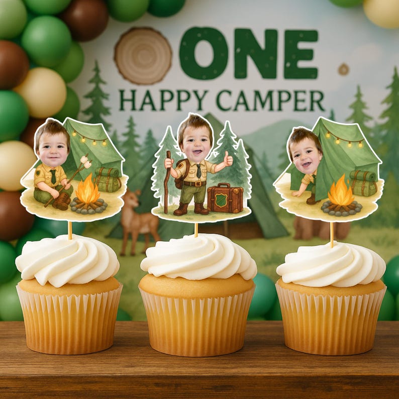 May include: Three cupcakes with white frosting, each topped with a decorative cutout featuring a cartoon boy in a camping scene. The cutouts include a tent, campfire, and the text "ONE HAPPY CAMPER."