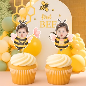 May include: Two cupcakes with yellow icing and paper cases. Each cupcake has a bee-themed topper featuring a baby's face. The background includes yellow and green balloons, and a sign that says "first BEE".