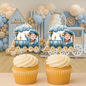 May include: Two cupcakes topped with blue train-shaped paper decorations featuring a child's face in the conductor's cabin. The train has gold wheels, a smokestack with blue smoke, and a gold bell. The cupcakes have white frosting in paper liners.