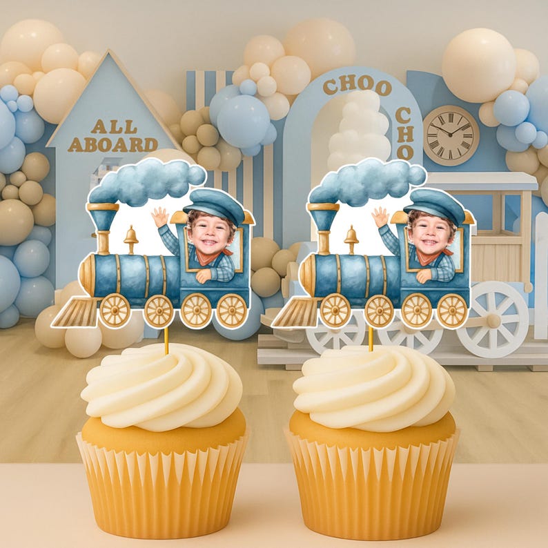 May include: Two cupcakes topped with blue train-shaped paper decorations featuring a child's face in the conductor's cabin. The train has gold wheels, a smokestack with blue smoke, and a gold bell. The cupcakes have white frosting in paper liners.