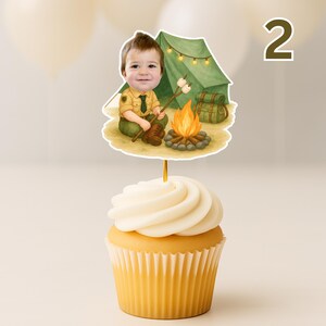 May include: A cupcake with white frosting topped with a decorative image of a young boy dressed as a scout, roasting marshmallows over a campfire. A green tent and backpack are in the background. The number "2" is in the background.