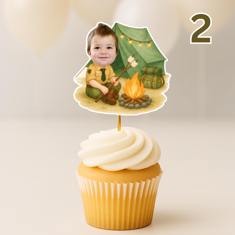 May include: A cupcake with white frosting topped with a decorative image of a young boy dressed as a scout, roasting marshmallows over a campfire. A green tent and backpack are in the background. The number "2" is in the background.