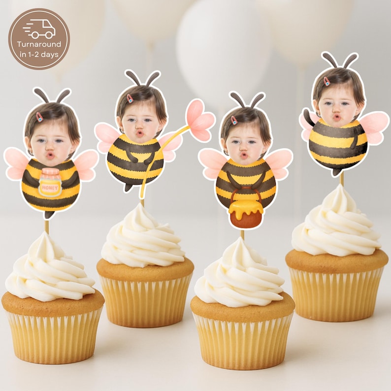 May include: Four cupcakes with white frosting and bee-themed toppers. The toppers show a child's face in a bee costume, holding honey pots and flowers. The cupcakes are on a white surface, with balloons in the background.