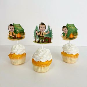 May include: Three cupcakes with white frosting and yellow cake. Each cupcake has a decorative topper featuring a cartoon boy in a scout uniform, a tent, and a campfire. The toppers are on white sticks.