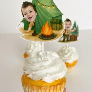 May include: Cupcakes topped with white frosting, featuring camping-themed toppers. The toppers depict a child in a camping scene with a tent, campfire, and backpack. The cupcakes have yellow cake and are in paper liners.