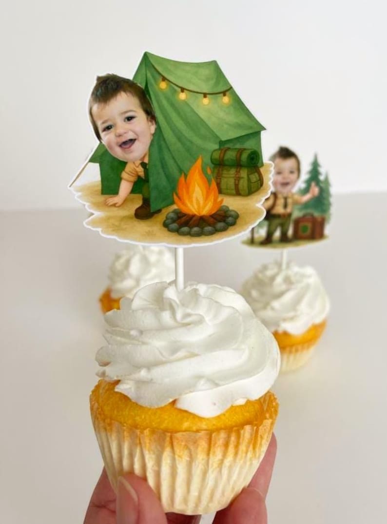 May include: Cupcakes topped with white frosting, featuring camping-themed toppers. The toppers depict a child in a camping scene with a tent, campfire, and backpack. The cupcakes have yellow cake and are in paper liners.