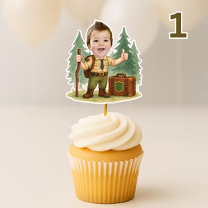May include: A cupcake topped with a decorative topper featuring a cartoon illustration of a child dressed as a park ranger. The child is holding a walking stick and giving a thumbs up. A brown suitcase is next to the child. The number "1" is in the background.