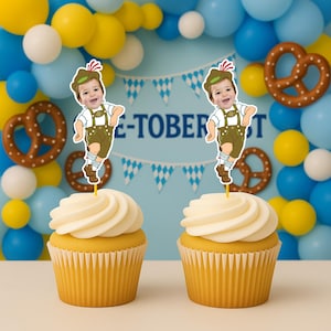 May include: Two cupcakes topped with yellow frosting and paper toppers featuring a cartoon illustration of a child wearing a green alpine hat, white shirt, and brown lederhosen. The toppers are positioned vertically on thin yellow sticks.