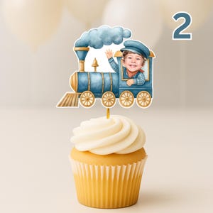 May include: A cupcake topped with a blue train cake topper featuring a watercolor-style illustration of a steam engine with a child's face in the conductor's cabin. The train has gold wheels, a blue smoke cloud, and a gold cowcatcher at the front.