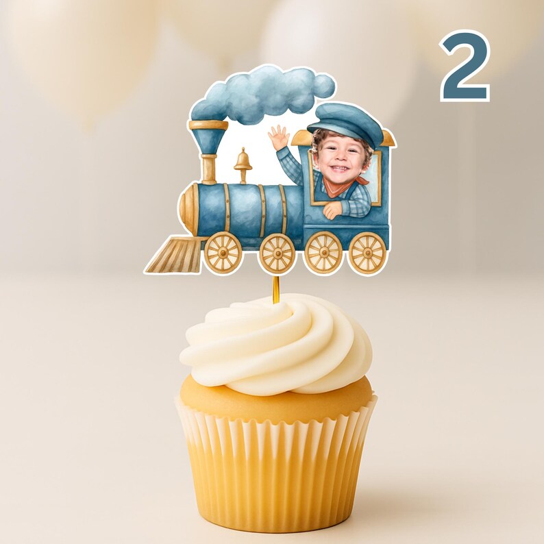 May include: A cupcake topped with a blue train cake topper featuring a watercolor-style illustration of a steam engine with a child's face in the conductor's cabin. The train has gold wheels, a blue smoke cloud, and a gold cowcatcher at the front.