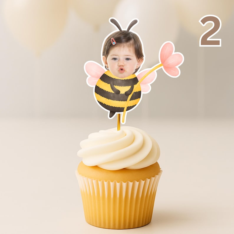 May include: A cupcake with a yellow paper case and white icing, decorated with a bee-themed topper featuring a child's face. The topper includes a yellow and black striped bee body, pink wings, and a flower.