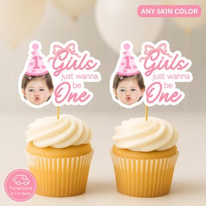 May include: Two cupcakes with white frosting and toppers that read "Girls just wanna be One." The toppers feature a photo of a baby wearing a pink party hat. The cupcakes sit in yellow paper cups.