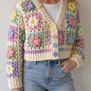 Könnte beinhalten: Eine cremefarbene, gehäkelte Strickjacke mit V-Ausschnitt und Knopfverschluss. Der Pullover hat bunte Blumenquadrate in Rosa, Gelb, Blau und Lila. Die Strickjacke wird mit hellblauen Jeans kombiniert.