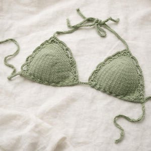 May include: A sage green crochet bikini top with triangle cups and scalloped edges. The top has tie closures at the neck and back. The handmade swimwear is displayed on a white, textured surface.