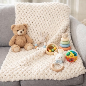 May include: A cream-colored, chunky knit baby blanket with a teddy bear, a bunny-shaped teether, a wooden rattle, a colorful stacking toy, and a multi-colored ball. The toys are arranged on the blanket, which is draped over a gray sofa.