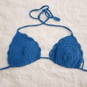 Könnte beinhalten: Handgefertigtes, blaues Häkel-Bikinioberteil. Das Oberteil hat einen gewellten Rand und Bindebänder am Hals und am Rücken. Die Dreiecks-Cups sind durch einen dünnen Häkelfaden verbunden. Das Bikinioberteil wird auf einer weißen Oberfläche präsentiert.