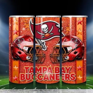 May include: A tumbler featuring the Tampa Bay Buccaneers logo, a red pirate flag with a skull and crossed swords, and two red football helmets. The words "TAMPA BAY BUCCANEERS" are displayed in red. The background is orange with stars.