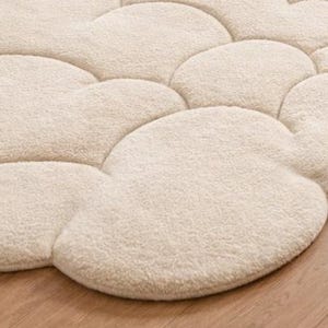 Puede incluir: Una alfombra crema con forma de nube con una superficie suave y texturizada. La alfombra est&aacute; formada por m&uacute;ltiples formas de nube interconectadas, creando una cubierta de suelo afelpada y acogedora. La alfombra est&aacute; colocada sobre un suelo de madera.