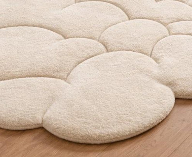 Puede incluir: Una alfombra crema con forma de nube con una superficie suave y texturizada. La alfombra est&aacute; formada por m&uacute;ltiples formas de nube interconectadas, creando una cubierta de suelo afelpada y acogedora. La alfombra est&aacute; colocada sobre un suelo de madera.