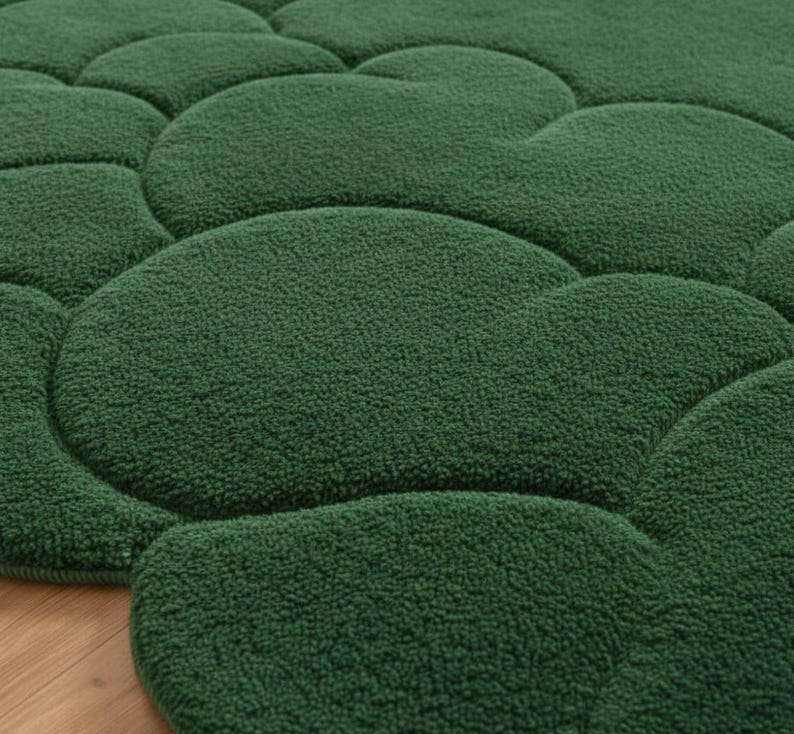 Puede incluir: Primer plano de una alfombra verde texturizada con un dise&ntilde;o &uacute;nico en forma de guijarros. La superficie de la alfombra es de un verde bosque profundo, con una textura suave y afelpada. El dise&ntilde;o presenta formas redondeadas e interconectadas, creando un patr&oacute;n visualmente interesante.