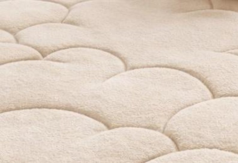 Puede incluir: Primer plano de un protector de colch&oacute;n acolchado beige con un patr&oacute;n de nubes. La tela tiene un aspecto suave y texturizado. Las costuras crean una serie de formas redondeadas e interconectadas.
