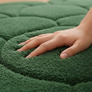 Puede incluir: Primer plano de una alfombra verde oscuro con un dise&ntilde;o texturizado y afelpado. La alfombra tiene un aspecto suave y acogedor, con una mano apoyada en su superficie, destacando su calidad t&aacute;ctil. El color de la alfombra es un verde bosque profundo.