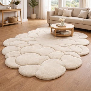 Puede incluir: Una alfombra en forma de nube de color crema con una superficie afelpada y texturizada. La alfombra est&aacute; compuesta por m&uacute;ltiples secciones redondeadas, creando una est&eacute;tica suave y acogedora. La alfombra est&aacute; colocada sobre un suelo de madera en un sal&oacute;n.