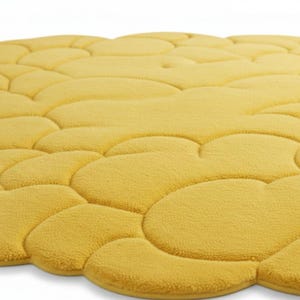 Puede incluir: Primer plano de una alfombra de ba&ntilde;o amarilla con un dise&ntilde;o de guijarros. La alfombra est&aacute; hecha de un material suave y afelpado y tiene una superficie texturizada. El color es un amarillo c&aacute;lido y soleado.