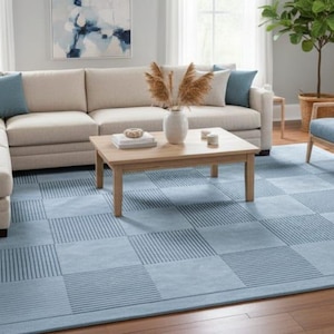 May include: A light blue area rug with a geometric pattern in a living room setting. The rug is placed under a beige sectional sofa, two blue armchairs, and a wooden coffee table. The room has a neutral color palette.
