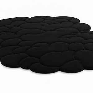 Puede incluir: Una alfombra negra con forma de nube y superficie texturizada. La alfombra est&aacute; formada por numerosas formas redondeadas e interconectadas, creando una apariencia suave y acolchada. La alfombra est&aacute; sobre un fondo blanco liso.