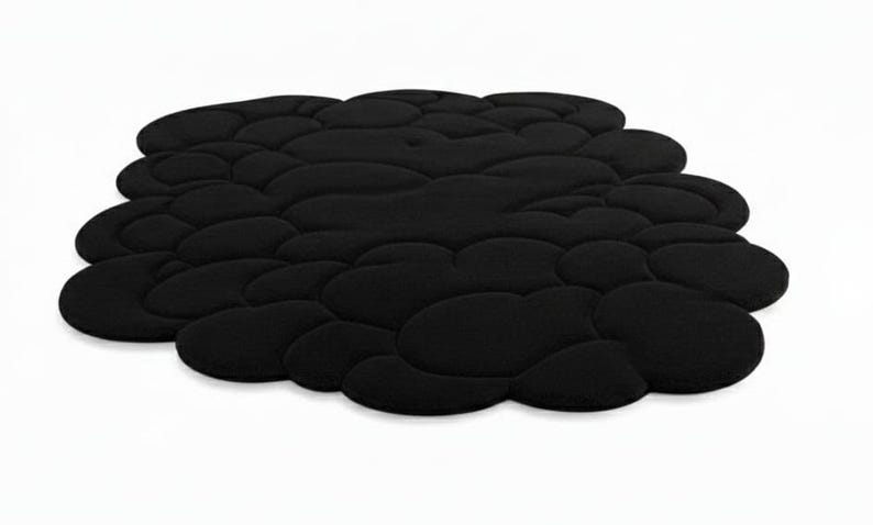Puede incluir: Una alfombra negra con forma de nube y superficie texturizada. La alfombra est&aacute; formada por numerosas formas redondeadas e interconectadas, creando una apariencia suave y acolchada. La alfombra est&aacute; sobre un fondo blanco liso.