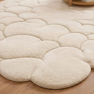Puede incluir: Una alfombra crema con forma de nube con una superficie suave y texturizada. La alfombra presenta un patr&oacute;n de formas redondeadas interconectadas, creando una apariencia afelpada y acogedora. La alfombra est&aacute; colocada sobre un suelo de madera.