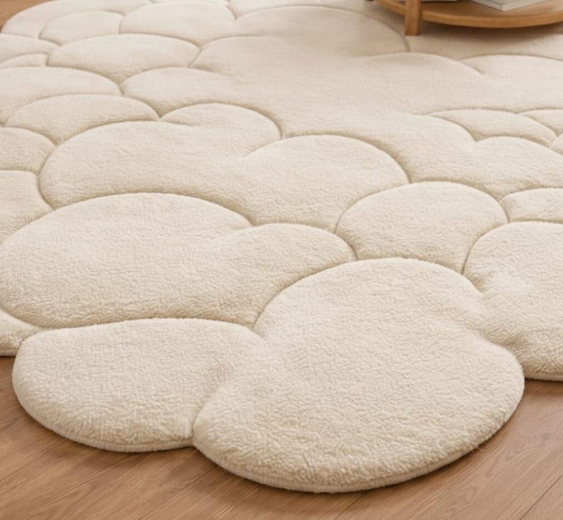 Puede incluir: Una alfombra crema con forma de nube con una superficie suave y texturizada. La alfombra presenta un patr&oacute;n de formas redondeadas interconectadas, creando una apariencia afelpada y acogedora. La alfombra est&aacute; colocada sobre un suelo de madera.