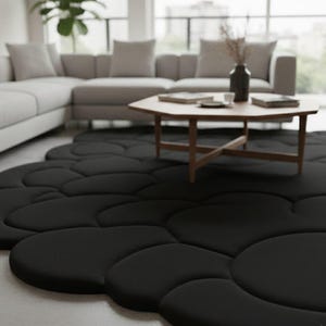 Puede incluir: Una alfombra negra con forma de nube y superficie suave y texturizada. La alfombra est&aacute; colocada sobre un suelo de color claro en un sal&oacute;n. Una mesa de centro de madera y un sof&aacute; seccional gris est&aacute;n en el fondo.