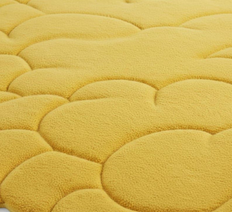 Puede incluir: Primer plano de una alfombra de ba&ntilde;o amarilla texturizada con un dise&ntilde;o similar a guijarros. La alfombra tiene un aspecto suave y afelpado y un color uniforme. El dise&ntilde;o presenta formas redondeadas e interconectadas.