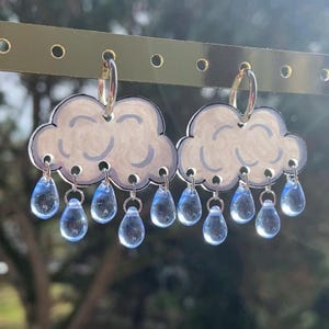 Può includere: Orecchini a forma di nuvola bianca con dettagli azzurri e gocce trasparenti a forma di lacrima. Sono appesi a cerchietti argentati. Un accessorio stravagante e originale.