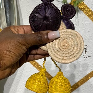 May include: Handmade earrings with a woven design. The earrings have a dark purple top, a beige circular middle, and bright yellow tassel-like bottoms. The earrings are held up by a silver hook.