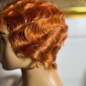 May include: A mannequin head with a short, wavy, orange-red wig. The wig has a vintage, finger wave style. The hair is styled close to the head, with defined waves. The mannequin's skin tone is light, and the background is blurred.