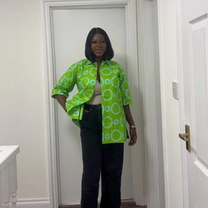 May include: A green button-down shirt with a light blue circle pattern. The shirt is paired with black pants. The outfit is worn with black sneakers. The shirt has three-quarter sleeves.