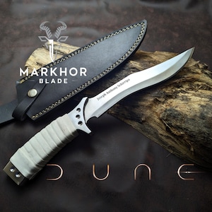 May include: A silver blade knife with a light gray wrapped handle and a black leather sheath. The knife has the text "Joseph Anthony Schirripa" engraved on the blade. The knife is displayed on a piece of wood with the word "DUNE" in copper-colored wire.