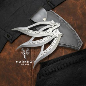 Puede incluir: Tres cuchillos arrojadizos plateados con hojas curvas y diseños de símbolos de infinito, sobre una funda de cuero y una superficie de cuero marrón. Los cuchillos tienen mangos decorativos y son de Markhor Blade.