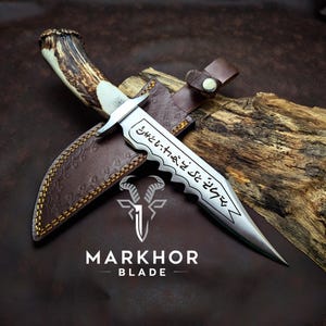 May include: A handcrafted dagger with a polished silver blade and a textured antler handle. The blade features etched text and a serrated edge. It rests in a brown leather sheath with decorative stitching. The Markhor Blade logo is visible.
