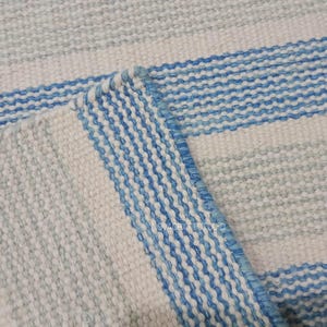 Puede incluir: Primer plano de una alfombra tejida con un patrón de rayas en tonos azules y blancos. La alfombra tiene una superficie texturizada y una esquina doblada, mostrando el tejido detallado. El diseño es sencillo y adecuado para la decoración del hogar.