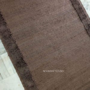 May include: A rectangular brown rug with a darker brown border. The rug has a textured appearance and is likely made of natural fibers. The text "NOVACRAFTSTUDIO" is visible on the rug.