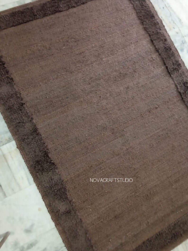 May include: A rectangular brown rug with a darker brown border. The rug has a textured appearance and is likely made of natural fibers. The text "NOVACRAFTSTUDIO" is visible on the rug.